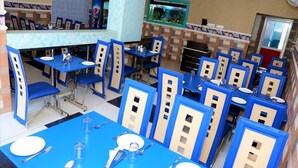 Um restaurante com assentos em azul e bege, mesas dispostas com pratos e talheres brancos, e um anúncio de Pepsi na parede.