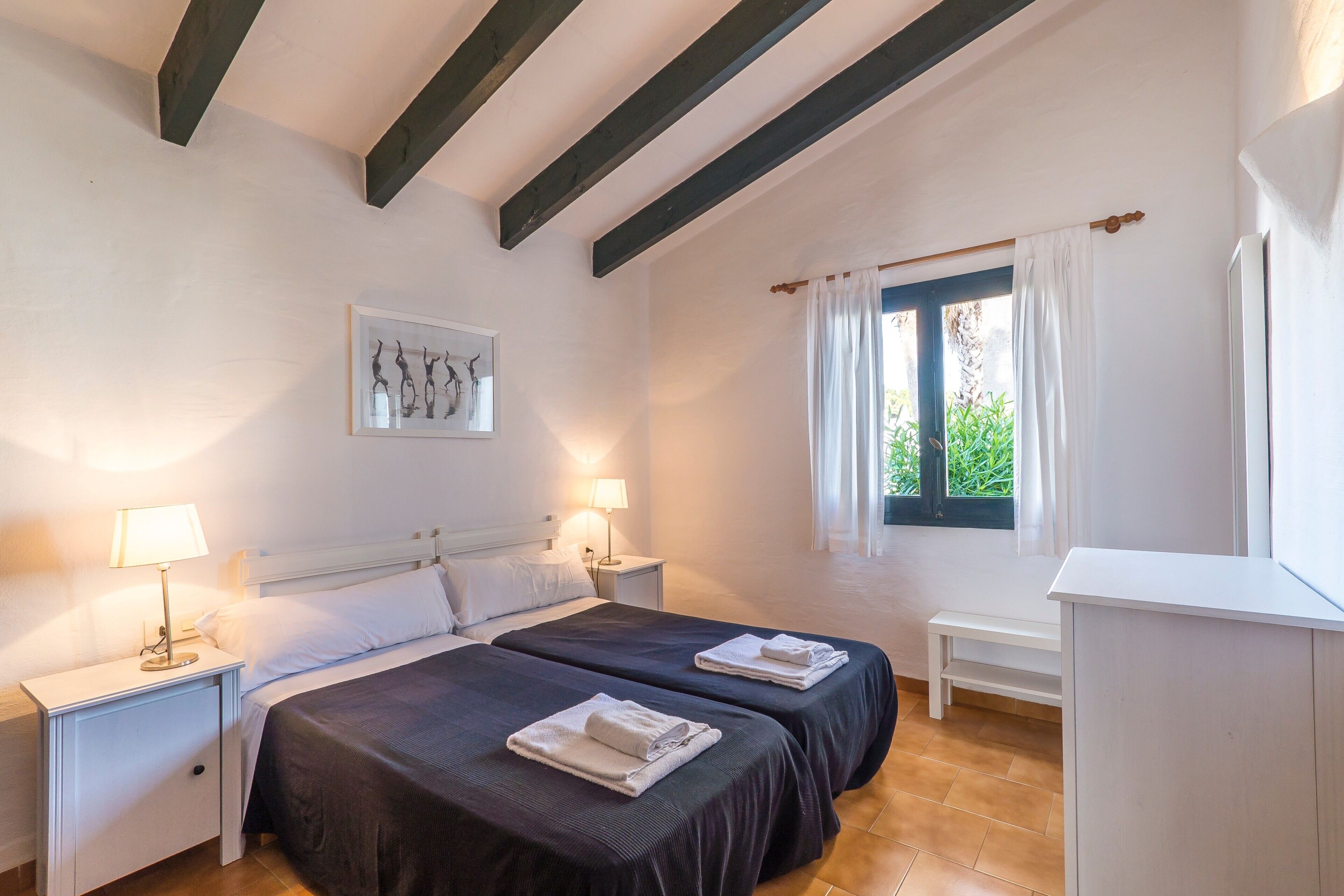 Un dormitorio con dos camas, una mesita de noche con lámpara, una ventana con cortinas y un escritorio blanco.