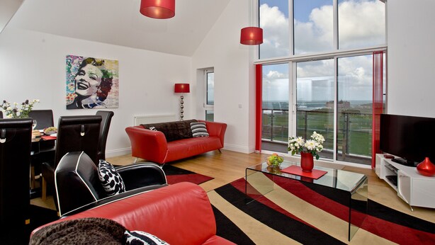 A modern living room with a red sofa, black and white patterned chairs, a glass coffee table, and a large window offering a city view. - Bredon Court, Poseidon Penthouse (Newquay)