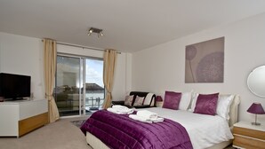 A modern hotel room with a large bed, a flat-screen TV, a sliding glass door to a balcony, and a view of the cityscape. - Bredon Court, Poseidon Penthouse (Newquay)