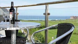 A balcony with a table, two chairs, a bottle of wine, and glasses. The view includes a grassy area and a building in the distance. - Bredon Court, Poseidon Penthouse (Newquay)