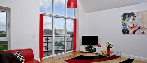 A modern living room with a red sofa, a glass coffee table, and a large window with a view of buildings.