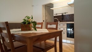 Ein Essbereich mit Holztisch und Stühlen, einer Topfpflanze auf dem Tisch und einer Küche im Hintergrund.
