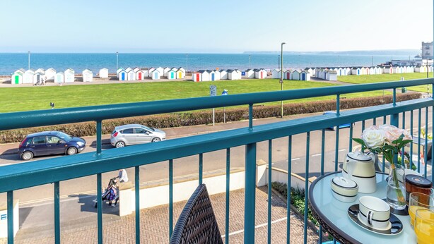 A balcony with a view of the sea, a row of colorful beach huts, and a table with breakfast items. - Comfort and luxury in a sea front location with panoramic sea views. (Paignton)