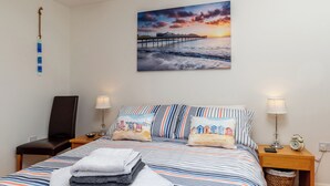 A neatly made bed with striped bedding, a wall-mounted painting of a sunset over a pier, bedside lamps, and a small wooden nightstand with a lamp and an alarm clock. - Comfort and luxury in a sea front location with panoramic sea views. (Paignton)