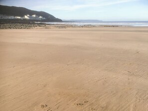 Une vaste plage de sable parsemée d’empreintes, une côte rocheuse et des bâtiments visibles au loin.