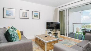 A modern living room with a grey sofa, a wooden coffee table, and a flat-screen TV. There are three framed pictures on the wall, a sliding glass door leading to a balcony, and a small table with a chair outside.