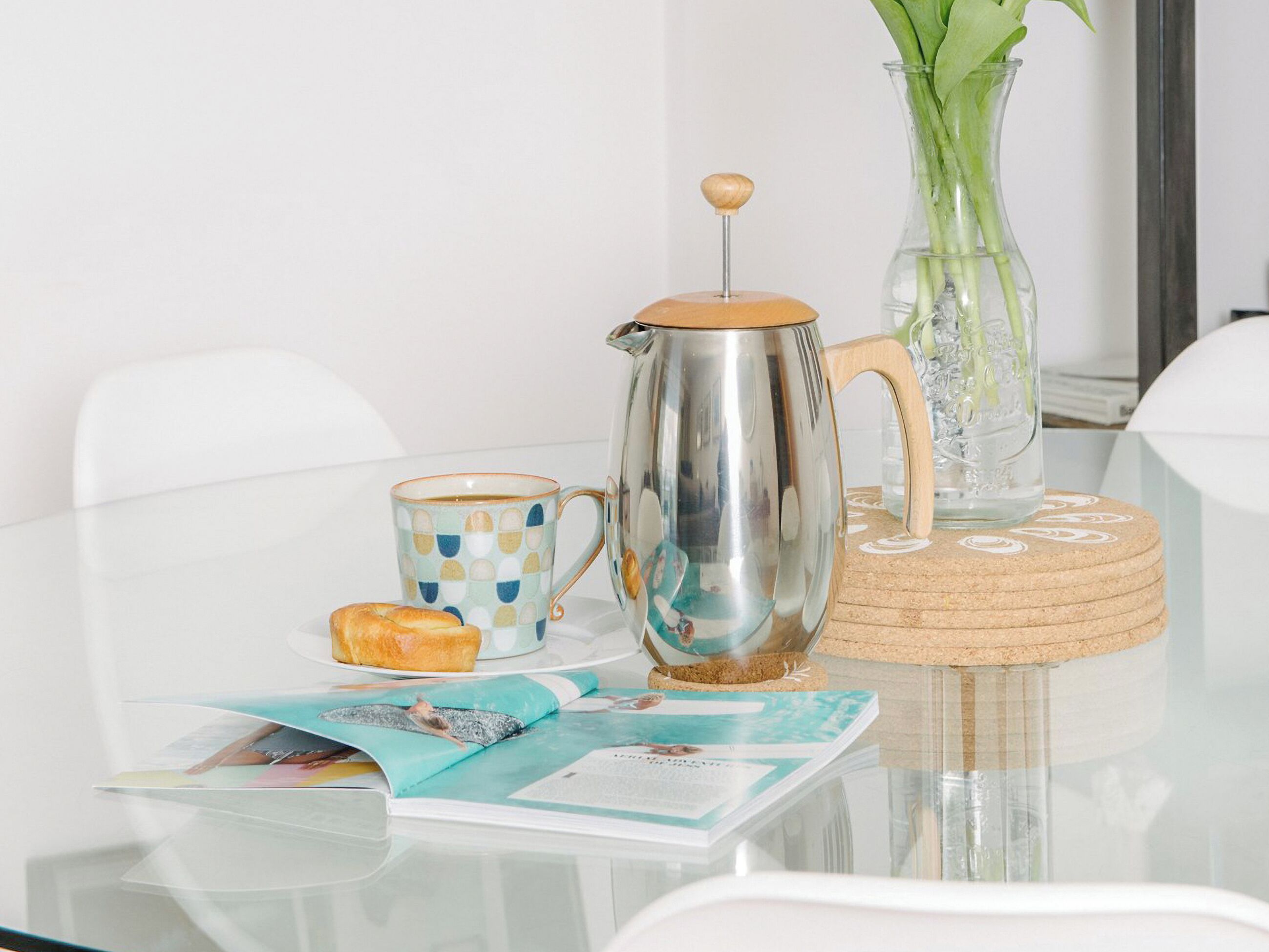 Ein Tisch mit einer Kaffeekanne, einer Tasse, einer Vase mit Blumen und einer Zeitschrift.