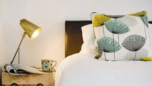 A neatly made bed with a white comforter and a pillow with a leaf pattern. A bedside table holds a lamp, a mug, and a magazine.