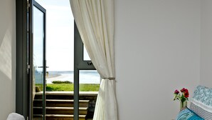 Une chambre avec vue sur la plage, un lit recouvert d’une couvre-lit à motifs bleus et blancs, un banc en bois à l’extérieur et un vase rempli de fleurs sur la table de chevet.