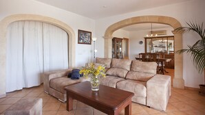 A living room with a beige sofa, a wooden coffee table, and a vase with yellow flowers. There is a dining area in the background with a wooden table and chairs. - Finca with sea views and pool special offer last week of August (Calonge)