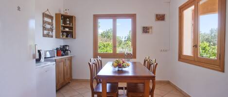A kitchen with a dining table, chairs, a coffee shelf, and a window with a view of greenery.