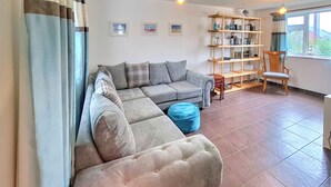 A living room with a sectional sofa, a blue ottoman, a wooden side table, a bookshelf, and a chair. - Trevistas, Padstow, Cornwall (Padstow)