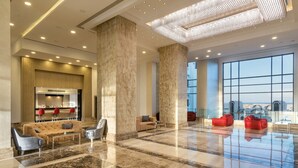Un hall d’entrée spacieux avec un sol en marbre, un coin salon doté de canapés et de fauteuils, un lustre moderne et de grandes fenêtres offrant une vue sur le paysage urbain.