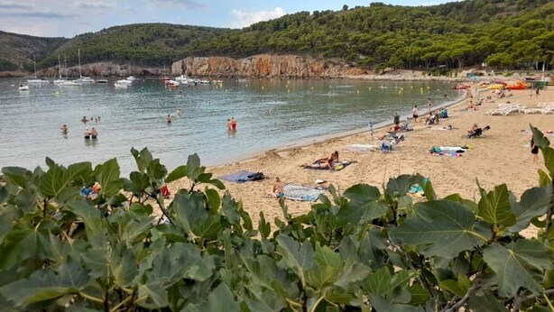 Una playa con gente nadando y tomando el sol, barcos en el agua y un paisaje montañoso al fondo.