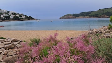 Una playa con un primer plano rocoso, mar tranquilo y edificios en una colina al fondo.