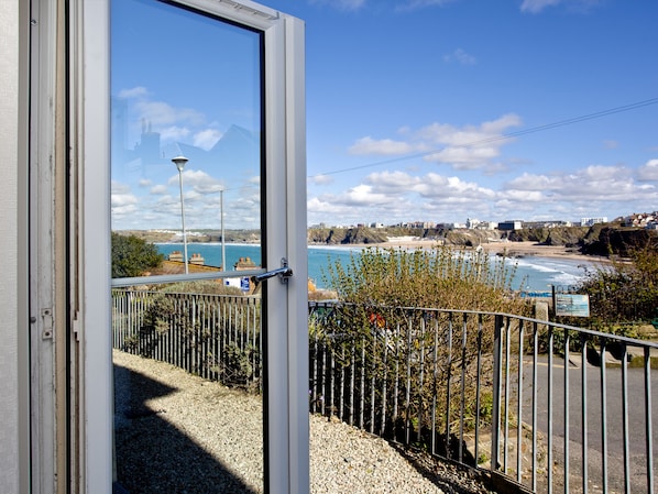 A coastal view through an open door, including a gravel path, metal fence, and distant buildings.