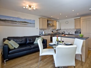 A modern kitchen with a dining area, featuring a black leather sofa, white chairs, and a glass table.