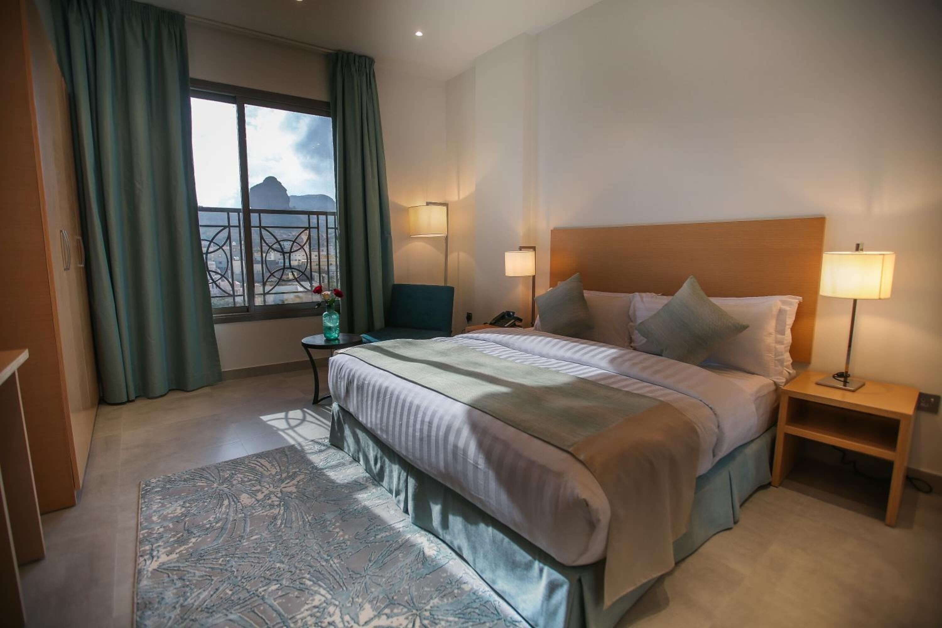 Et hotelværelse med en stor seng, to sengeborde med lamper, udsigt til et bjerg gennem et vindue og et lille bord med en vase.