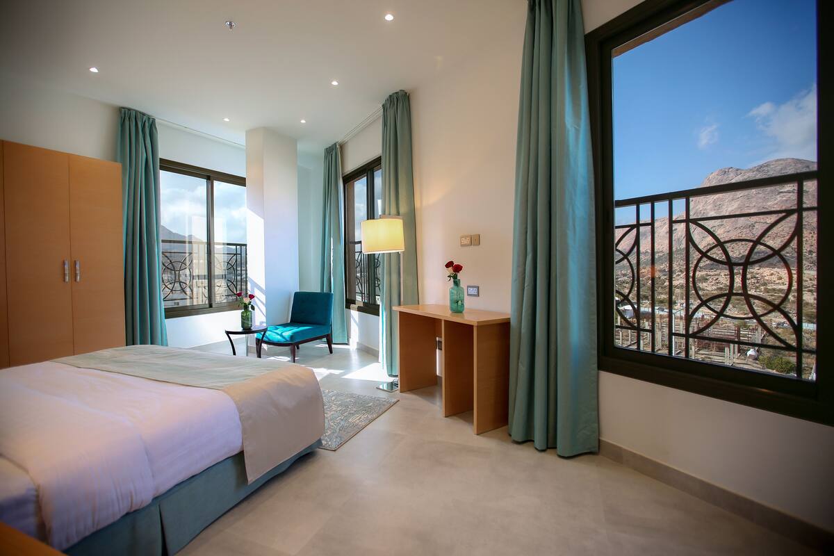 A modern hotel room with a large bed, a desk, a chair, and a view of a mountainous landscape through a window.