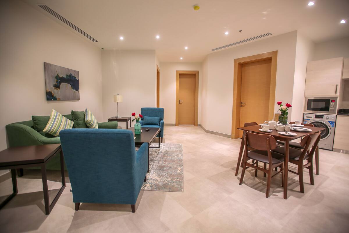 A modern living room with a green sofa, blue armchairs, a dining table, and a kitchen area with a microwave.