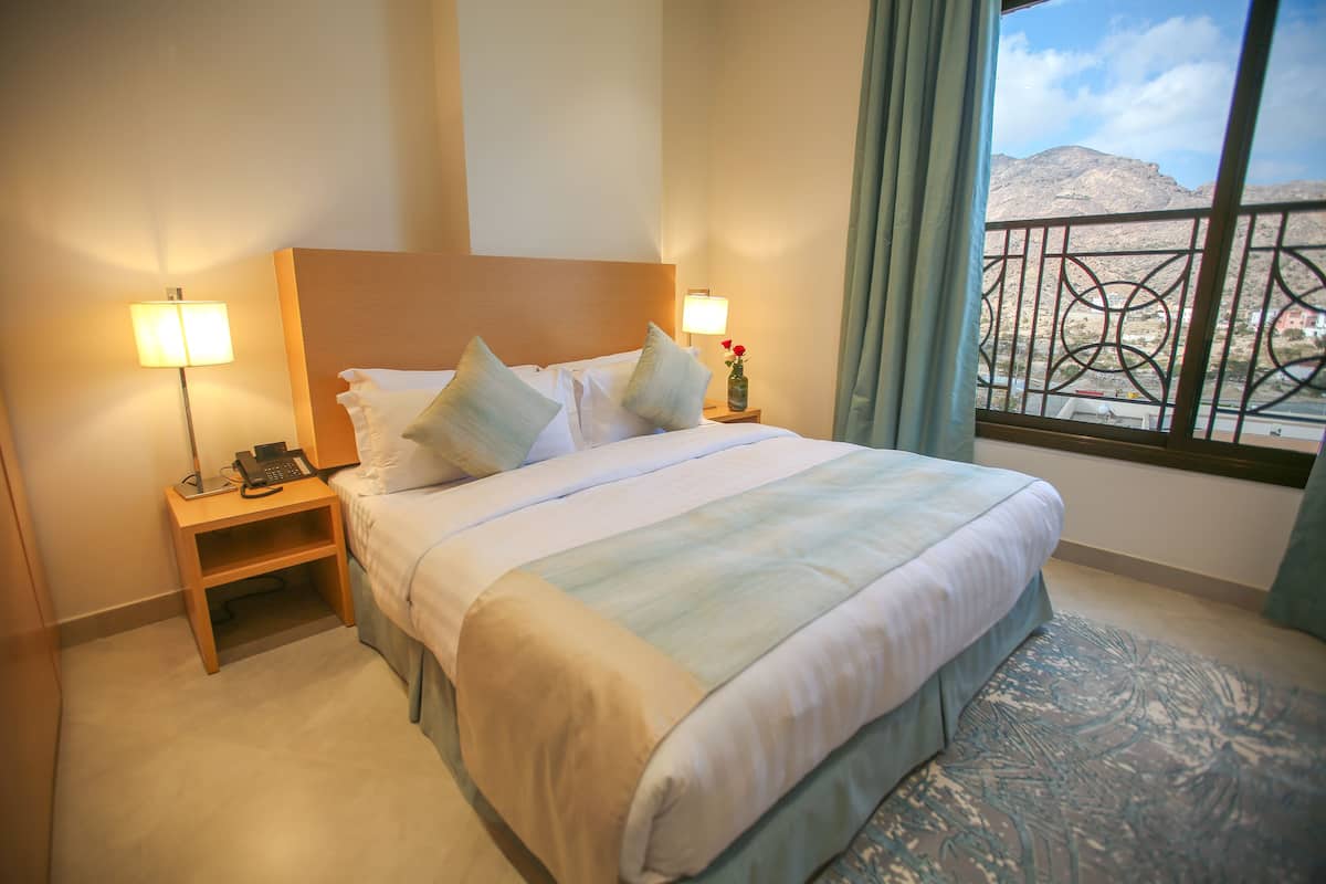 A hotel room with a bed, bedside lamps, a nightstand, a vase with a flower, and a view of mountains through a window.