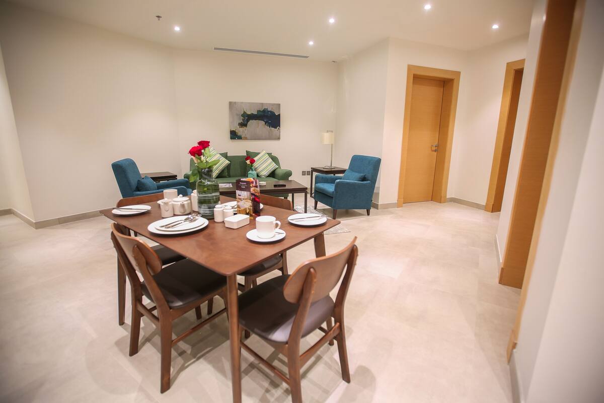 A dining area with a wooden table set for four, surrounded by blue chairs. There is a green sofa and a small side table with a lamp.
