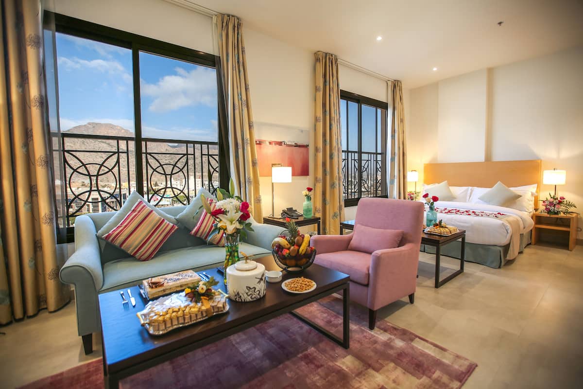 A hotel room with a balcony, a sofa, a chair, a dining table with food, a bed with a headboard, and a nightstand with a lamp.