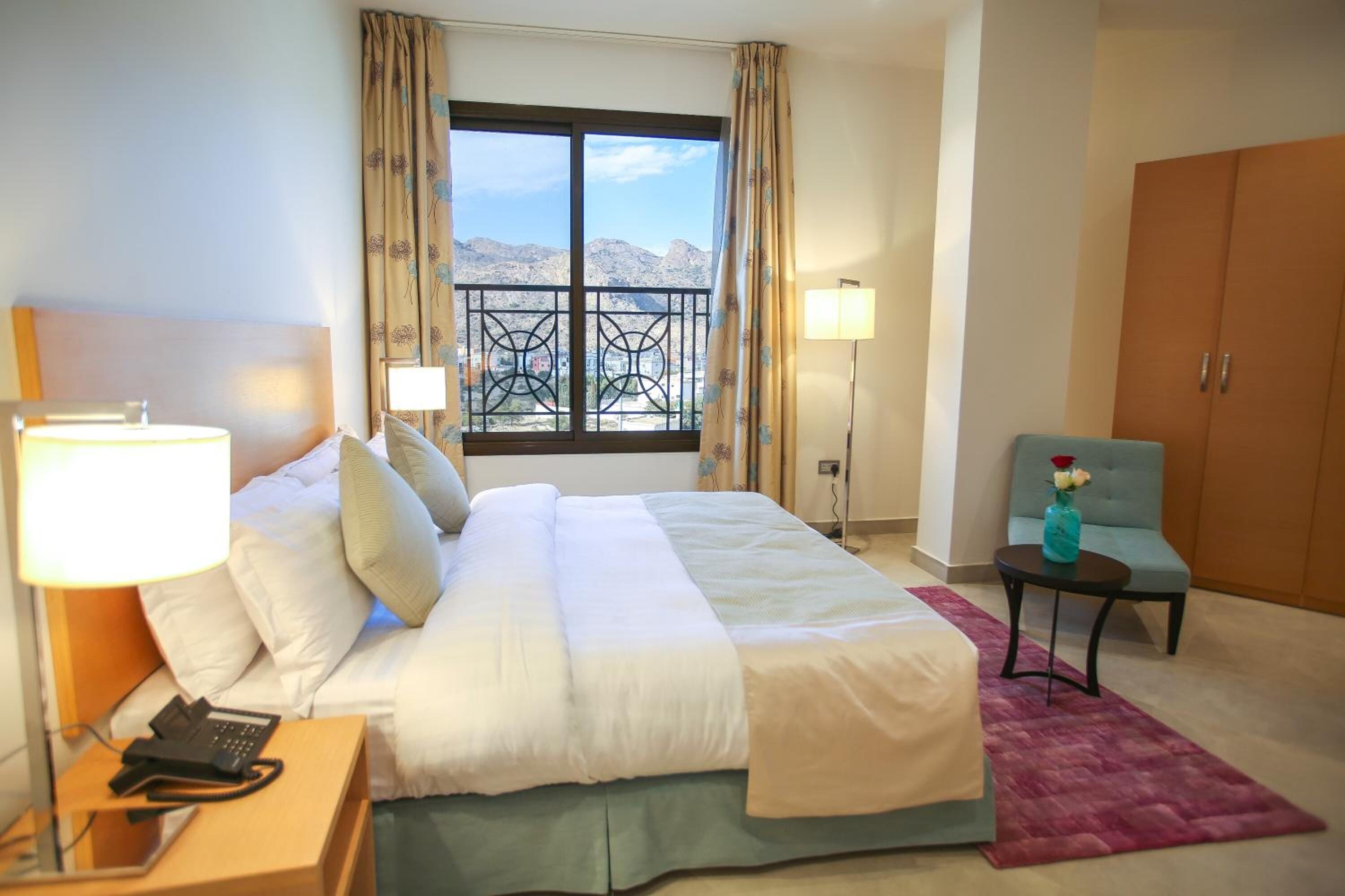 Quarto de hotel com uma cama, abajures, uma cadeira, uma escrivaninha com um telefone e uma janela com vista para montanhas.