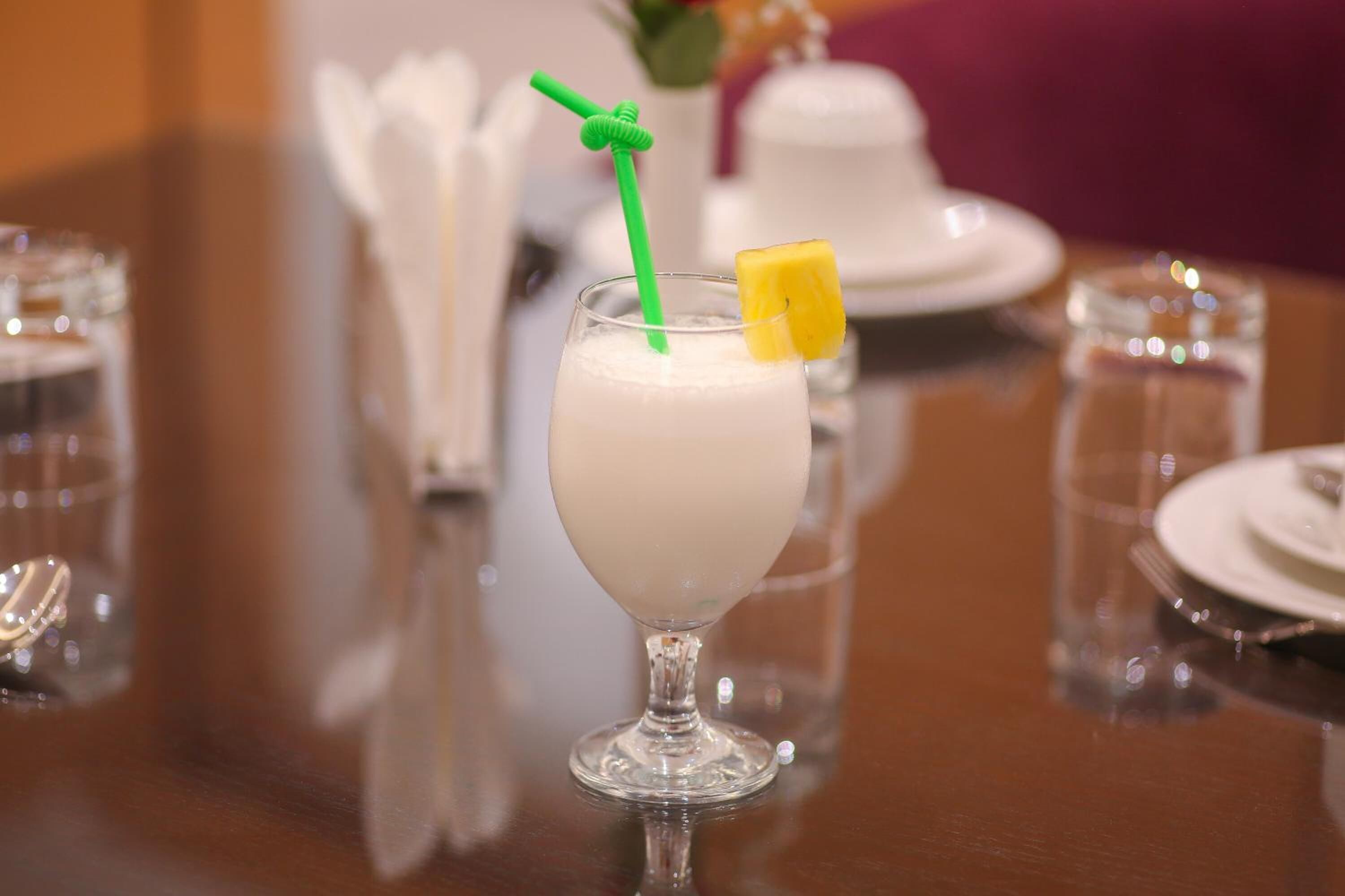 A glass of a creamy beverage with a green straw and a slice of fruit on the rim, placed on a reflective wooden table.