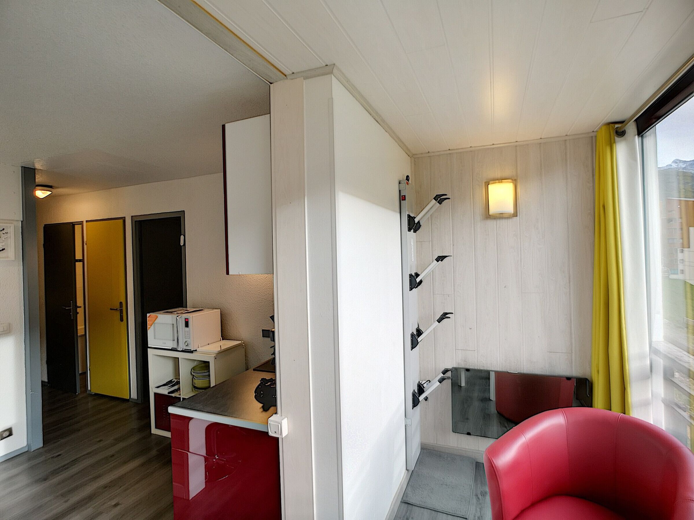 A compact hotel room with a kitchenette, a red chair, and a view through a window with yellow curtains.