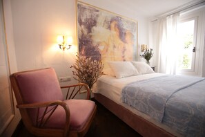 Un dormitorio con una cama, una silla, un cuadro y un florero con flores.