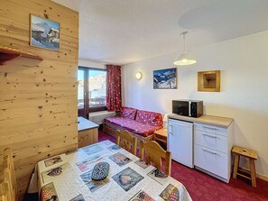 A cozy room with a wooden wall, a bed, a desk with a computer, a small table with a quilt, and a window with a view of buildings.