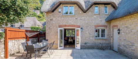Ein Steinhaus mit einem Strohdach, eine Terrasse mit Gartenmöbeln und eine Steinmauer.