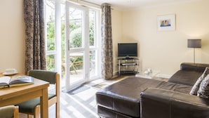 A modern living room with a leather sofa, a wooden dining table with chairs, a flat-screen TV, and a large window with curtains. - Longships Apartment - two bedroomed apartment within a 15 min walk of the beach. (Gwithian)