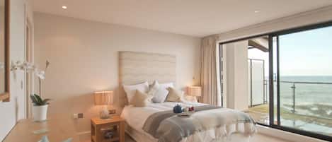 Une chambre avec un grand lit, des tables de chevet, une petite table de nuit, une plante en pot et une vue sur la mer depuis un balcon.