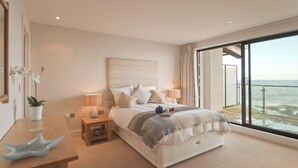 Een slaapkamer met een groot bed, nachtkastjes, een plant in pot en uitzicht op zee vanaf een balkon.