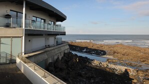 Een modern gebouw met twee verdiepingen en een balkon met uitzicht op de oceaan, een rotsachtige kustlijn en een heldere lucht.