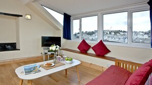En stue med et fjernsyn, et bord med et magasin, en skål med frugt og en rød sofa.