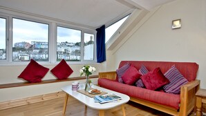 En stue med en rød sofa, et hvidt sofabord og udsigt til en kystby gennem store vinduer.