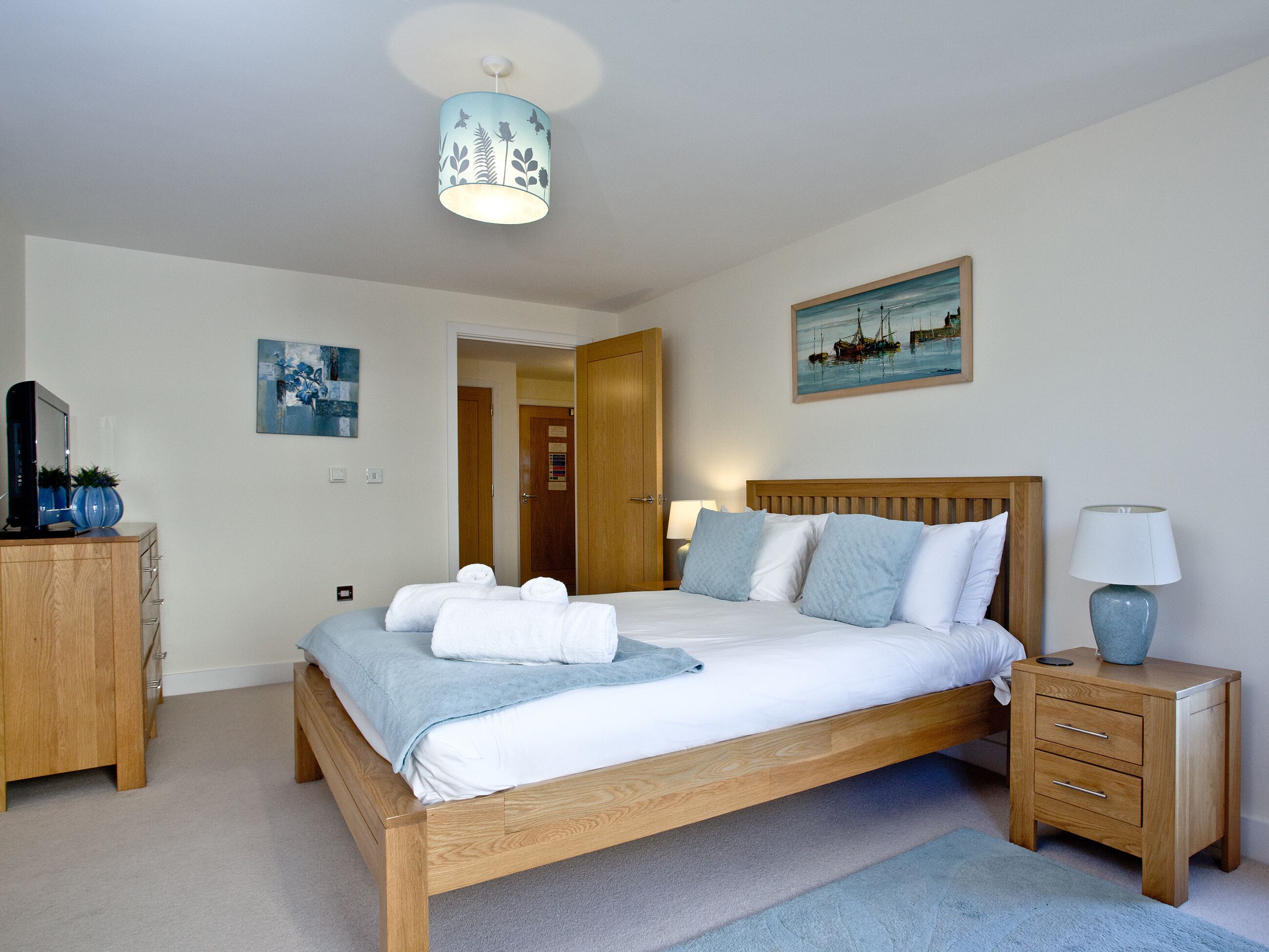 A neatly arranged bedroom with a wooden bed, bedside tables, a nightstand, a television, and artwork on the walls.