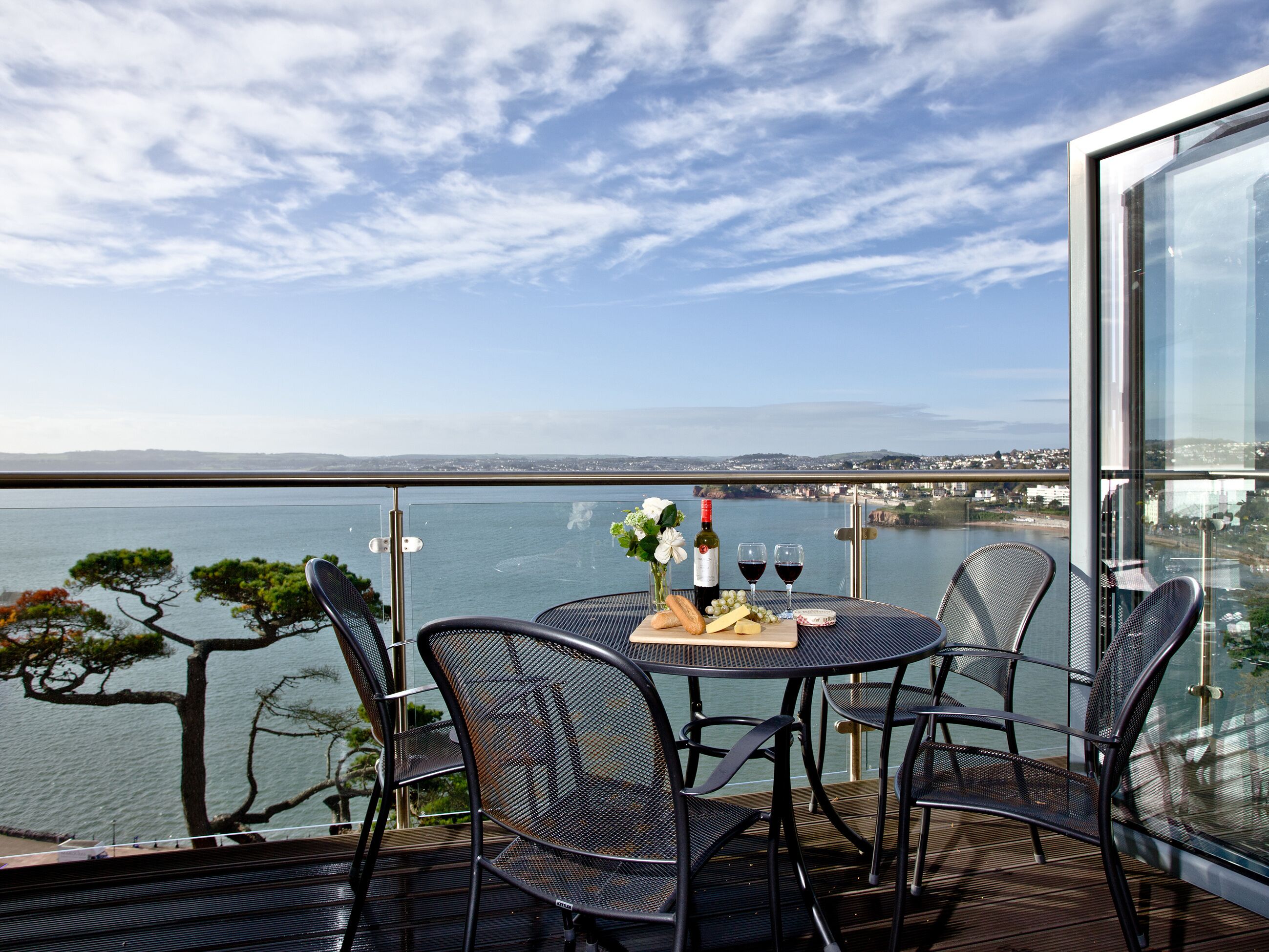 A balcony with a table set for two, overlooking a body of water and a distant cityscape.