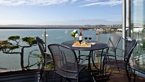 Un balcon avec une table dressée pour deux personnes, offrant une vue sur une étendue d’eau et sur les contours d’une ville lointaine.