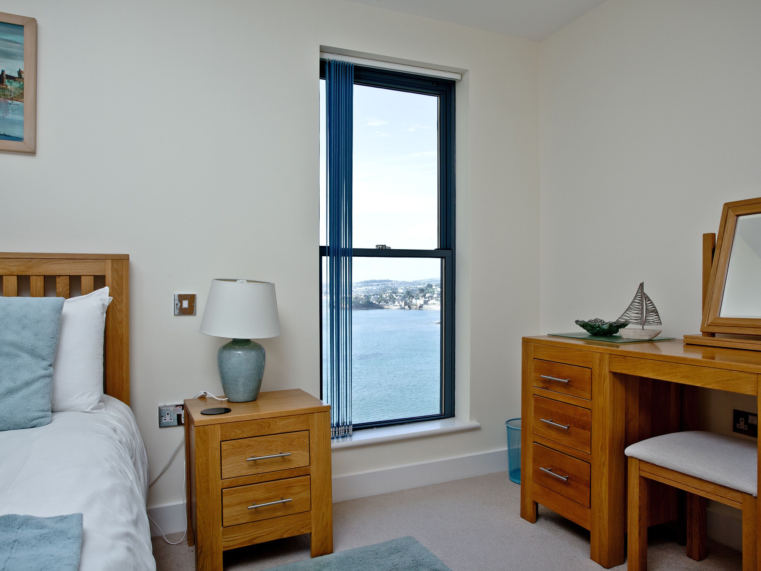 A bedroom with a bed, a nightstand, a desk, and a chair. There is a window with a view of a body of water and a distant landscape.
