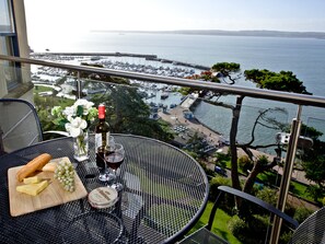 Ein Balkon mit Tisch für zwei Personen, Blick auf einen Yachthafen mit Booten und einer fernen Küstenlinie.