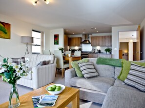 A modern living room with a sofa, armchairs, a coffee table with a bowl of apples, and a dining area with a table and chairs.