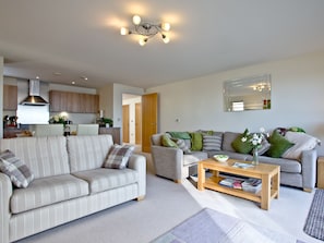A modern living room with a sofa, a coffee table, and a dining area with a kitchen in the background. - Harbreck Heights -Masts B9 (Torquay)