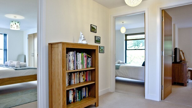 Une pièce avec une bibliothèque en bois remplie de livres, un lit recouvert d’une couette blanche et une fenêtre donnant sur un paysage verdoyant.