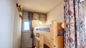 Una habitación pequeña y funcional con una cama nido, una mesita, una ventana con cortinas y un radiador.