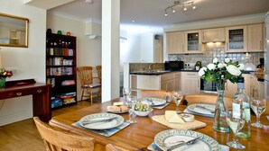 A dining area with a wooden table set for four, a bookshelf, and a kitchen in the background. - A spacious apartment overlooking Goodrington Park with sea views. (Paignton)